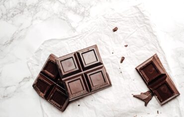 Broken dark chocolate bar pieces on white parchment paper and marble surface with scattered chocolate crumbs